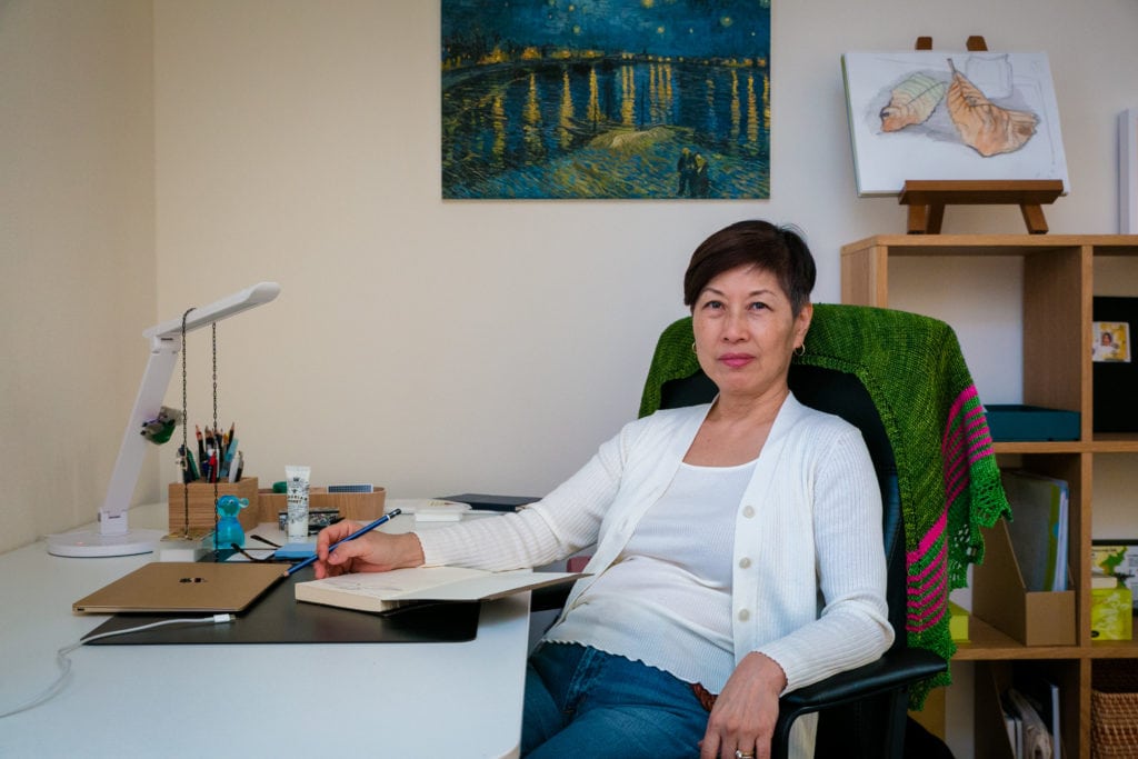 Liz Noh sitting at desk with book open, pencil in hand, looking at camera